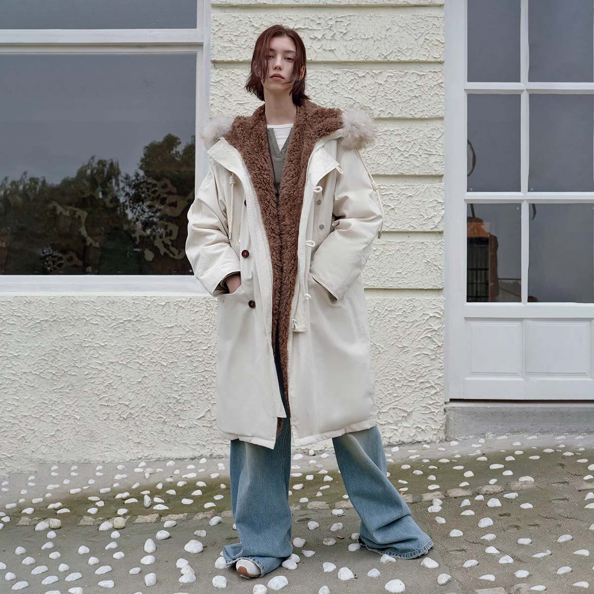 Person wearing a long cream coat with brown fur lining standing in front of a white building.
