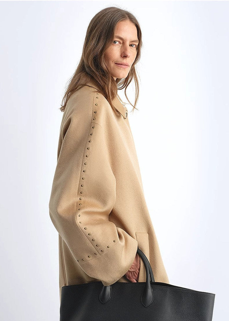 Woman wearing a beige coat with studded details, holding a black handbag on a white background
