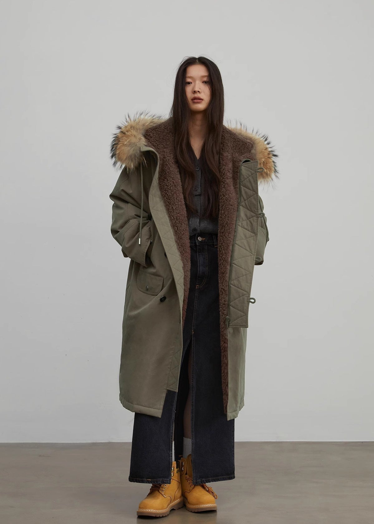 Person wearing a long olive green coat with fur trim against a plain background